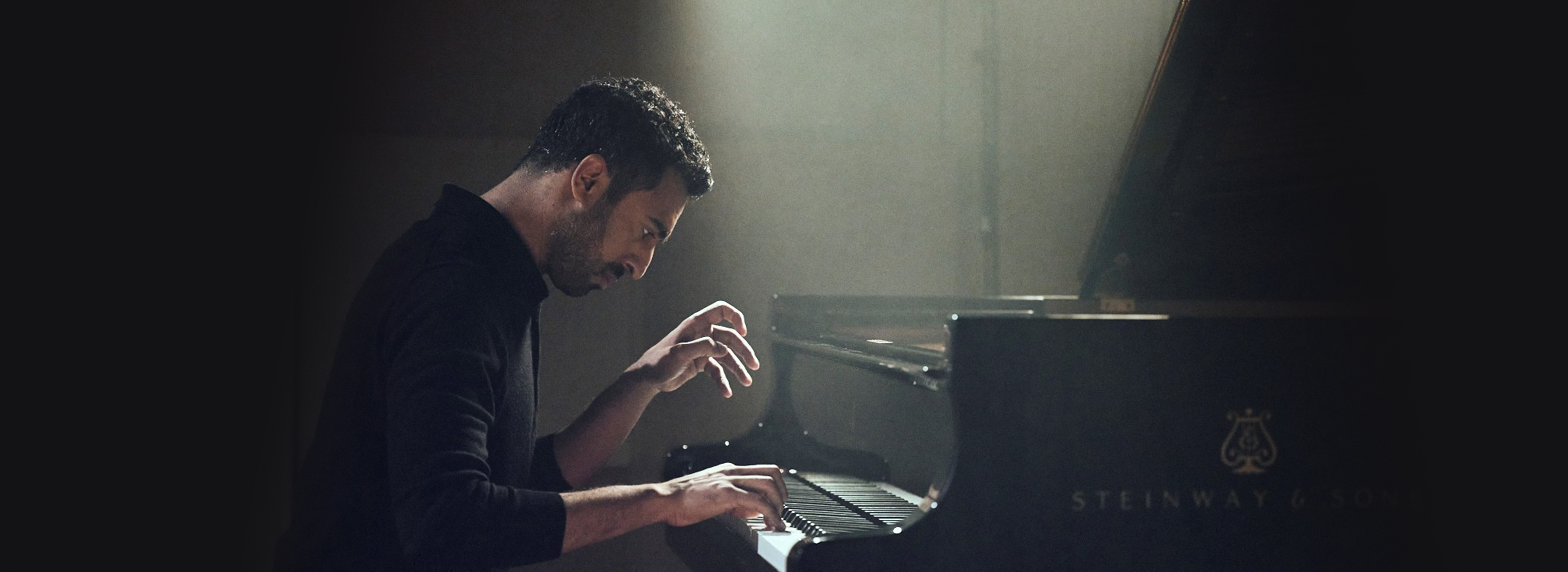 A man wearing a black jumper playing the piano under a spotlight.